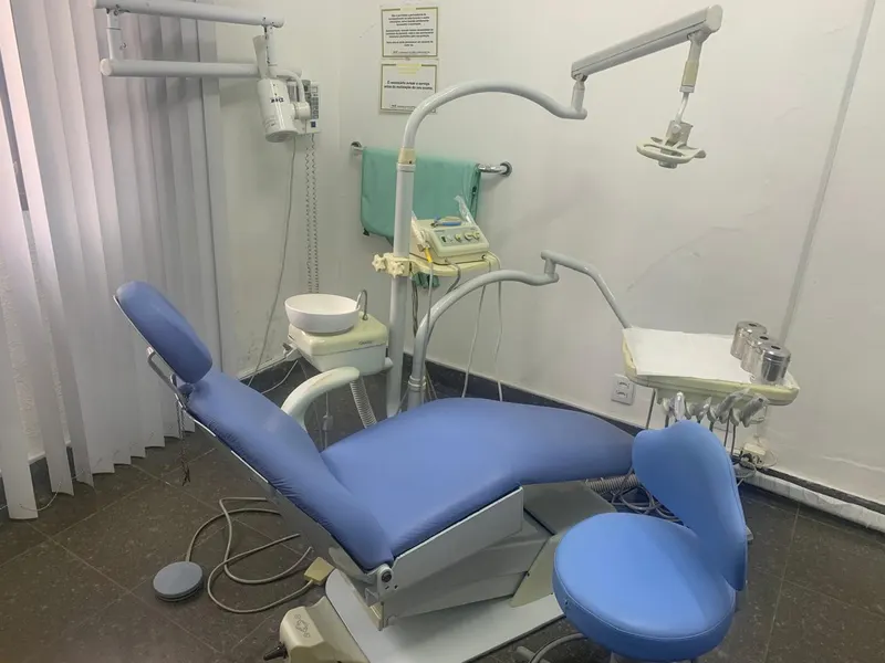 Consultório com cadeira odontológica azul-clara, refletor e equipamentos junto à parede branca.
