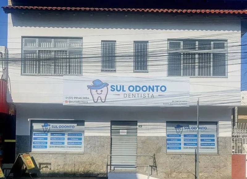 Fachada da Sul Odonto: sobrado branco com base de pedra cinza, a placa azul com o dente de chapéu e as janelas com os serviços listados.