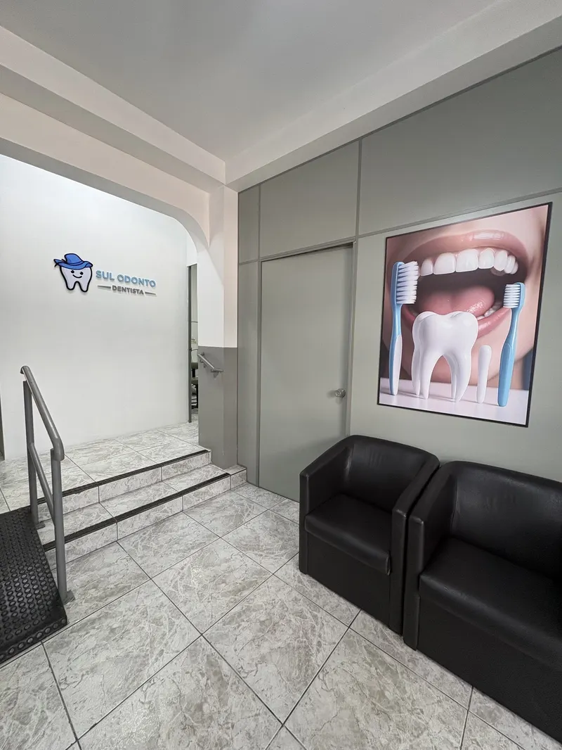 Recepção da Sul Odonto: escada com corrimão, dois sofás pretos e o logo azul na parede branca.